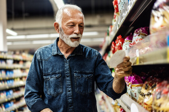 Mature male buyer shopping groceries in supermarket from shelf standing with shopping list in hand.