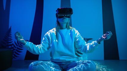 Girl wearing a virtual reality headset sitting in a lotus pose and meditating in a room with blue...