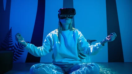 Fotobehang Girl wearing a virtual reality headset sitting in a lotus pose and meditating in a room with blue neon light and glowing neon lines. © 24K-Production