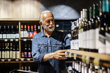 Male consumer buying bottle of red wine in winery store. Smiling mature man wearing casual clothes shopping at supermaket grocery store buy choosing wine alcohol hold bottle inside hypermarket.