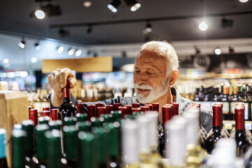 Positive male looking for perfect wine. Man choosing wine bottle picking up from alcohol shelf at wine store shop while he is looking at product and smile.