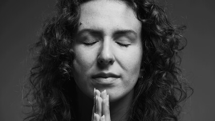 Spiritual 20s person feeling GRATITUDE and Godly devotion with eyes closed and hands clenched in PRAYER. Hopeful woman having FAITH in monochromatic, black and white