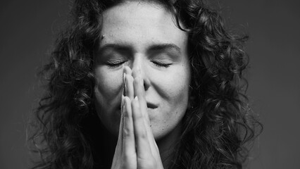 Spiritual 20s person feeling GRATITUDE and Godly devotion with eyes closed and hands clenched in PRAYER. Hopeful woman having FAITH in monochromatic, black and white