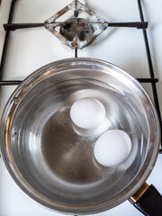 two white chicken eggs are boiling in steel ladle on gas stove close up