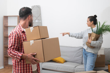 Young woman with packed house plant in hands points index finger to man with cardboard parcels to...