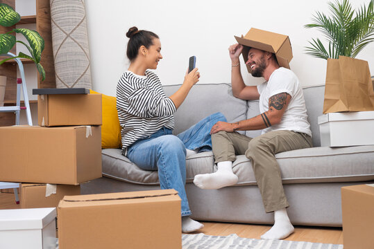 Young couple of newlyweds in love does packing things with furniture with carpets in numerous different cardboard moving boxes taking funny memorable photos in old home