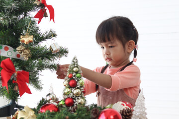 Cute little Asian girl child decorating beautiful Christmas tree with ornaments in white living room. Concentrate kid celebrating winter holiday. Merry Christmas and Happy New year. Happy childhood