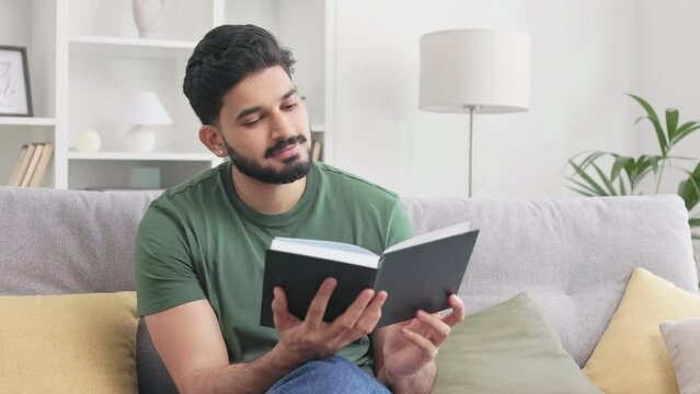 Portrait of male book lover opening new one literary work while relaxing on comfort sofa in personal apartments. Handsome indian man enjoying favorite reading genre indoors.