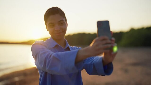 Teenager girl makes selfie on sea beach. Indian teen girl with short hair smiling looking at smartphone camera at sunset resting on ocean beach. Taking picture. photographing, social media addiction.