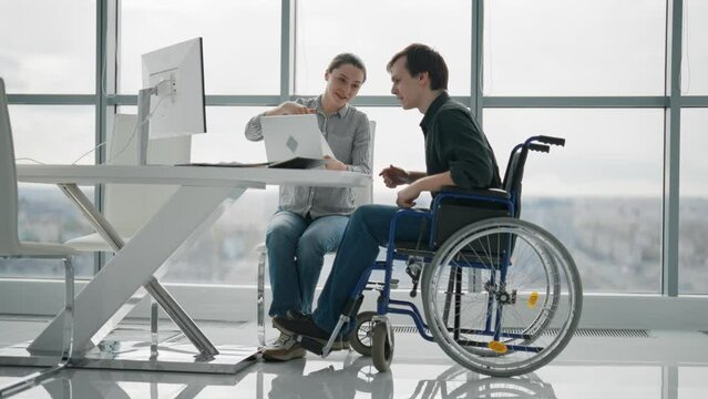Paralyzed man work with woman in office. Colleagues discussing project together female shows to guy in wheelchair pie chart on paper document. Positive workers discuss analytics data sit at desk.