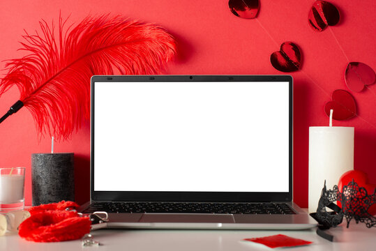 Provocative tableau featuring side view laptop, candles, mask, adult toys like furry handcuffs and feather tickler on red background. Explore online world with empty screen, a canvas for your desires