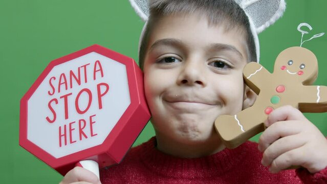 Cute boy with christmas sweater and deer alntlers holding sign Santa stop here and gingerbread man. Isolated on green background, slow motion close up. High quality 4k footage