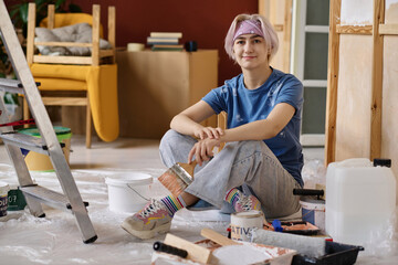 Portrait of young painter with brush sitting on the floor and looking at camera while making repair in the room