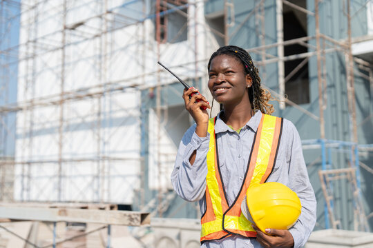 African American engineer woman using radio talking instructing and checking building progress on construction site. Confident female foreman standing in safety vest and working outside.