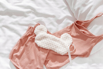 Pink female pajama and fluffy fur sleep eye mask on white bedclothes background. Top view comfort...