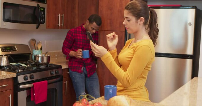 Caucasian woman using her cell phone in the morning with boyfriend drinking coffee in the background. African Millennial couple in the kitchen on mobile. 4k slow motion handheld