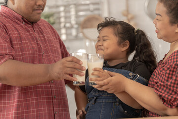 A chubby family with a father wearing a prosthetic. They were happily inviting their girl to drink morning milk together in the kitchen of their home. Breakfast time of Asian dad mom and kid people. - obrazy, fototapety, plakaty