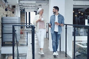 Smiling man ans woman holding coffee while walking in the office hall, talking, having coffee break. Full-length. Teamwork, startup, business concept - obrazy, fototapety, plakaty