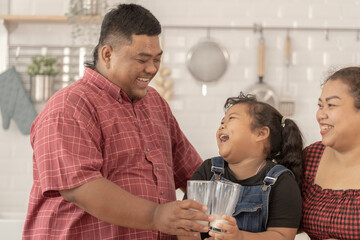 A chubby family with a father wearing a prosthetic. They were happily inviting their girl to drink morning milk together in the kitchen of their home. Breakfast time of Asian dad mom and kid people.