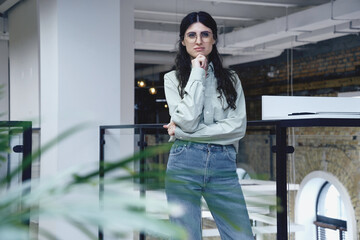 Young beautiful Caucasian female business worker with hand on chin standing at office. Startup, strategy, business concept
