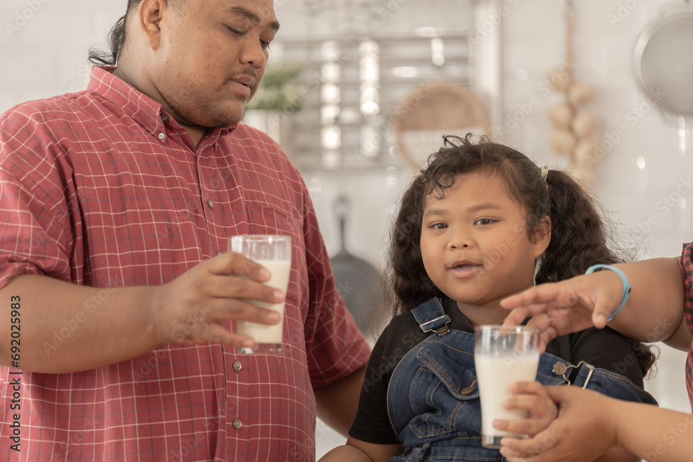 Wall mural A chubby family with a father wearing a prosthetic. They were happily inviting their girl to drink morning milk together in the kitchen of their home. Breakfast time of Asian dad mom and kid people.