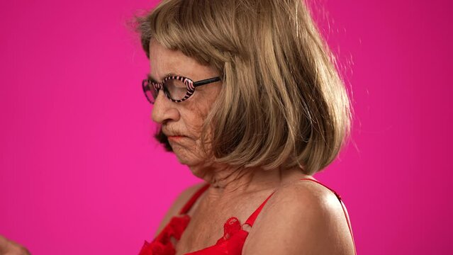 Closeup of funny elderly woman walks across frame while scrolling on her smart phone, stops smiles at camera and continues across frame. Isolated on pink background
