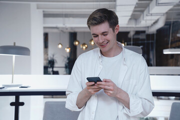 Smiling young man typing on the smartphone while standing in the office. Startup, strategy, business concept