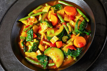 vegetable soup with green beans, broccoli, carrot, celery, spinach, cabbage, zucchini, tomatoes in black bowl on concrete table with spoon, vertical view, close-up