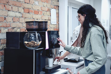 Young woman preparing fresh aromatic coffee with modern machine while standing in the office lobby. Teamwork, startup, business concept