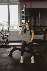 Fototapeta na wymiar Plump woman takes break from exercise on machine drinking water and using smartphone in sports club. Improving physical health