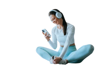 Zelfklevend Fotobehang Cheerful brunette American girl in sportswear, headphones sitting on floor against transparent background holds phone looks at screen reads message at break of exercise, happy the receive great news © Iona