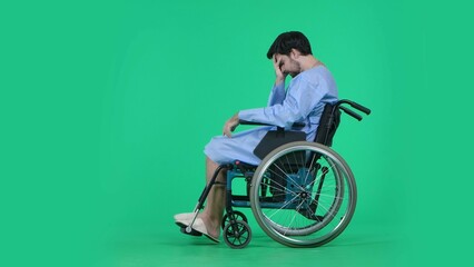 Adult man patient in robe sitting in wheelchair holding head in hand, looking at his legs,...
