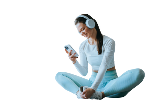 Cheerful brunette American girl in sportswear, headphones sitting on floor against transparent background holds phone looks at screen reads message at break of exercise, happy the receive great news