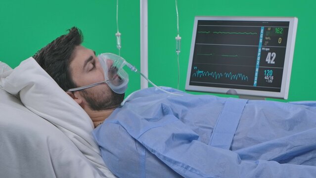 Adult man patient laying in bed with drip, breathing mask and monitor, eyes closed, heart rate blood pressure signs absent. Isolated on chroma key green screen.