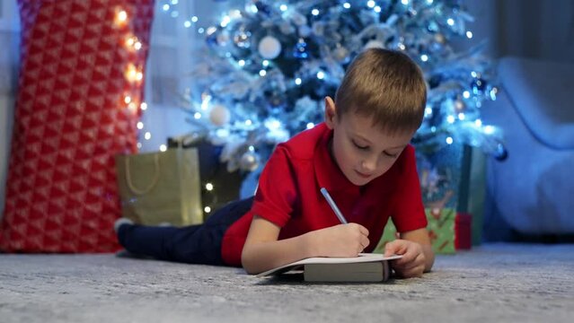 The boy writes a letter to Santa Claus near the Christmas tree. The child is preparing for the holiday and waiting for gifts. High quality 4k footage