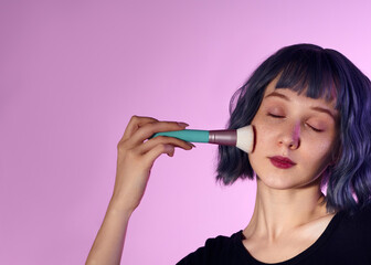 A cheerful woman in a blue wig with red freckles uses a fluffy makeup brush