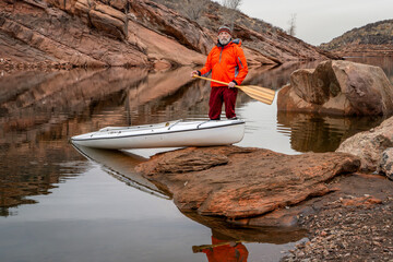 senior male paddler with a decked expedition canoe with a wooden paddle on a rocky shore of...