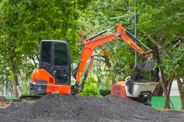 Orange mini excavator in operation digs and loads in the back of a mini stretcher bulldozer fertile soil in the park. - obrazy, fototapety, plakaty