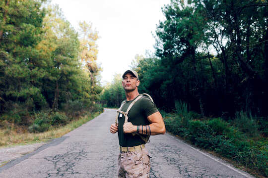 Army man in uniform with backpack and cap standing on road against trees in forest