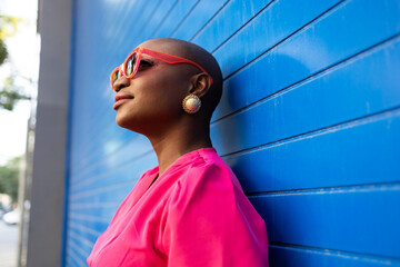 Happy Stylish black woman with shaved head in sunglasses and pink dress standing against blue wall