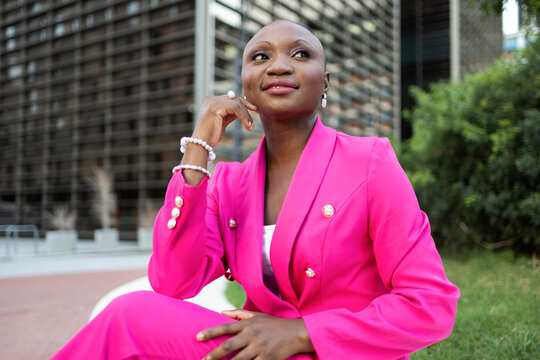 Smiling black lady with shaved head in trendy pink outfit sitting on street against modern building