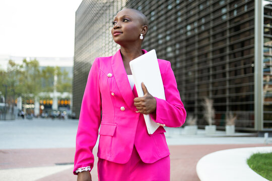 Elegant African American female entrepreneur with shaved head in pink outfit with computer walking on street