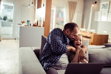 Happy little girl sitting on couch with father using laptop