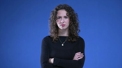 Upset woman with arms crossed looking at camera with angry expression, standing on blue background. Female person in 20s with negative body language