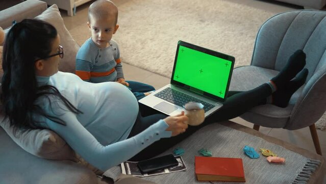 Pregnant mother with child using laptop with green screen at home