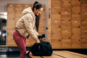 A fit sportswoman is packing sports clothes in her bag while standing in gym fitting room.