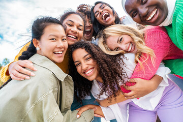 Multicultural group of young women smiling at camera outdoors - Happy girl friends having fun together hanging out on city street - Female community concept with girls taking selfie picture with phone