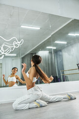 Fototapeta na wymiar artistic african american dancer in white sportswear exercising near mirror in studio, choreography