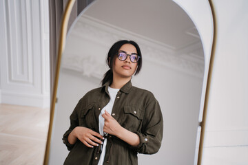 Young African American lady in casual shirt standing at mirror at living room looks away with...