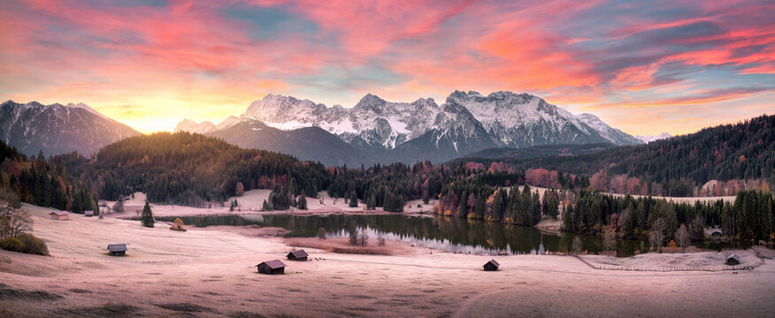 Stunning sunrise scenery with mountains, a scenic lake and woods on hills in the Alps, frozen meadow and cabins, beautiful nature panoramic shot